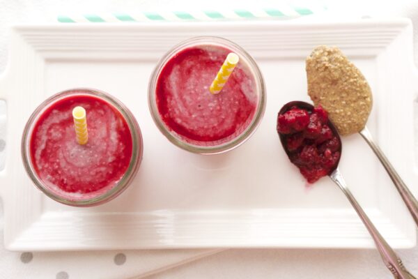 Top view of a two smoothies with a table spoon of raspberries and a tablespoon of almond butter on a white tray