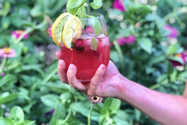 Hand holding Raspberry Basil Lemonade garnished with lemon and basil with flower background