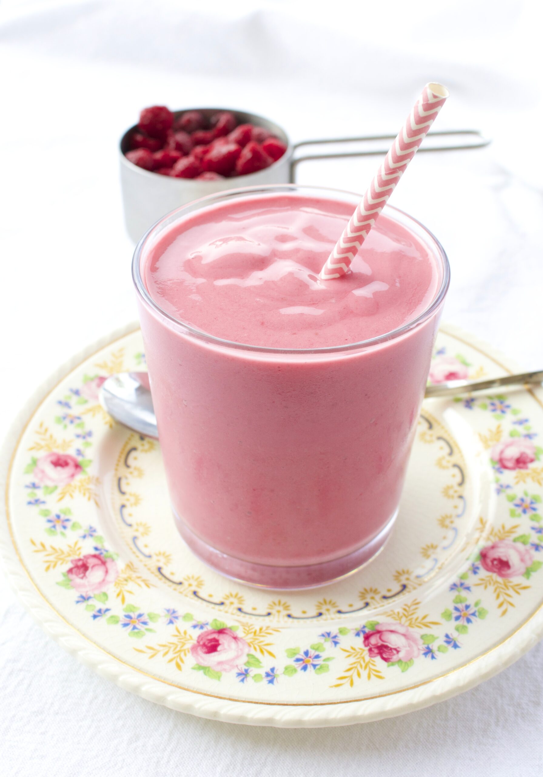 Raspberry Cheesecake Smoothie in short glass with a pink straw on a vintage plate decorated with flowers
