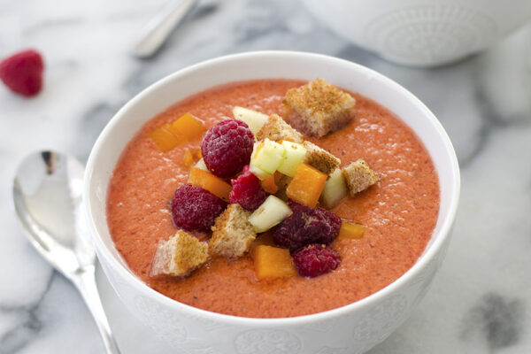 Two white bowls filled with Raspberry Gazpacho, garnished with raspberries, bread crumbs, bell pepper and cucumber on a marble table.